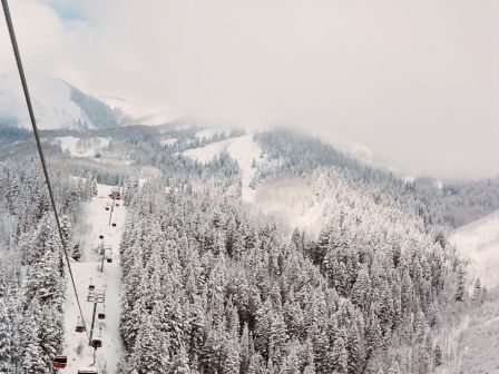 A snow-covered mountainous landscape with a series of ski lifts ascending the slope, surrounded by densely forested areas blanketed in snow.