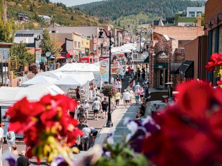 A bustling street scene with people walking, colorful flowers in the foreground, various shops, and buildings lining the street, against a mountainous backdrop.