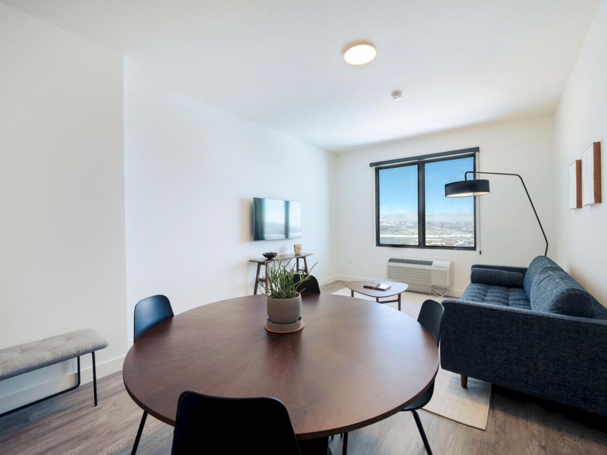 A modern living room with a round dining table and four chairs, a sofa, a wall-mounted TV, a window with a view, a floor lamp, and a bench.