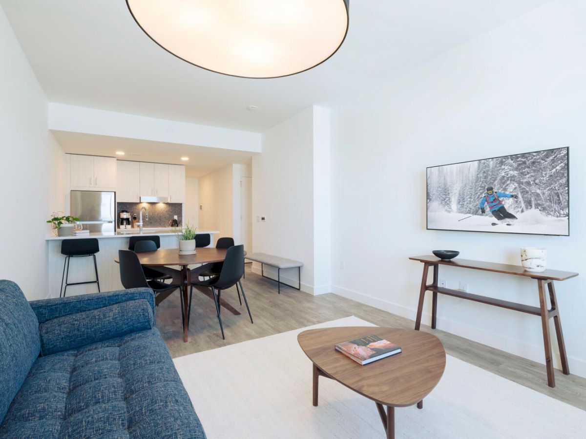 A modern, minimalist open-concept living and dining area with a blue sofa, wooden furniture, and a kitchen in the background.