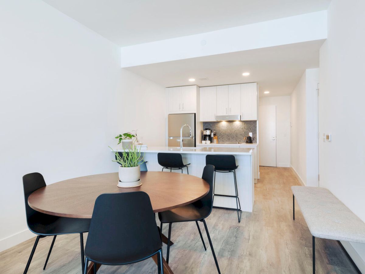 A modern kitchen and dining area with a round table, chairs, a potted plant, and bar stools at the kitchen counter, under bright lighting.