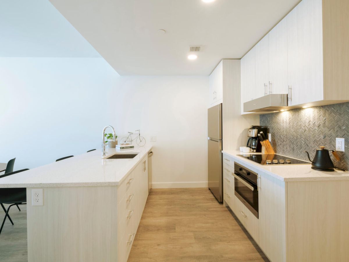 This image shows a modern kitchen with light wood cabinets, a stove, a microwave, a coffee maker, and a kettle, leading to a dining area.