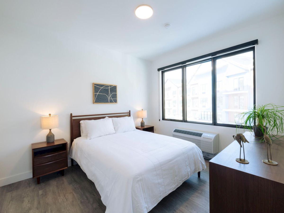 A modern bedroom with a bed, two bedside tables, lamps, a dresser, a window, and a wall art piece, all in a well-lit, minimalist setting.