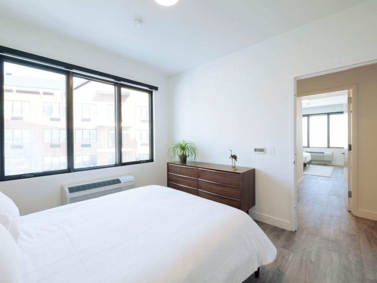 A modern, minimalist bedroom with a white bed, wooden dresser, large windows, plant, and doorway leading to an adjacent room with another window.