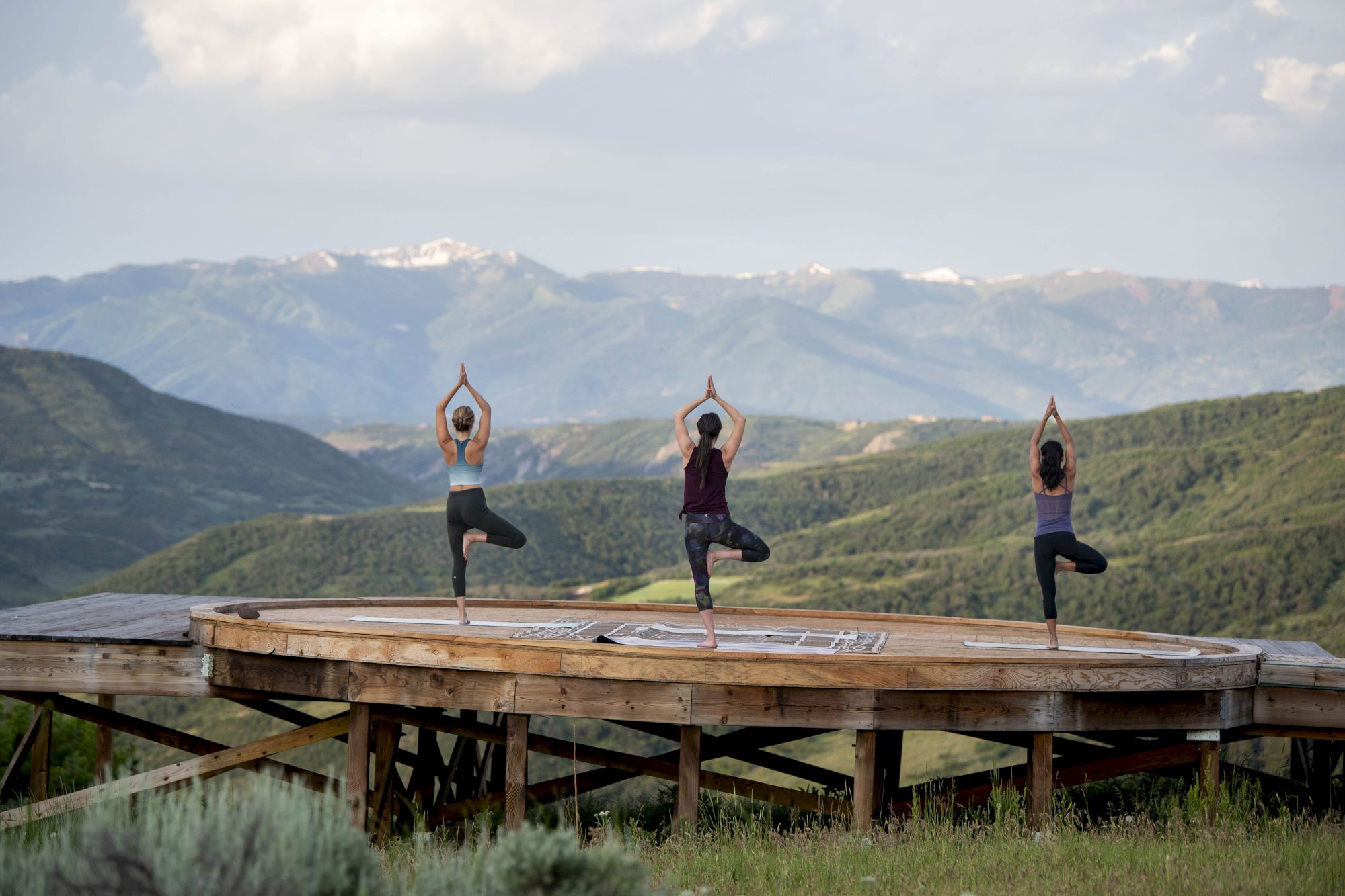 Three people practice tree pose (yoga) on a wooden platform overlooking green hills and distant snow-capped mountains.