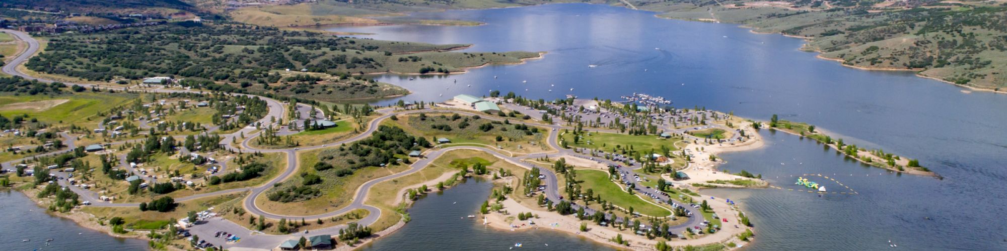 An aerial view of a lake surrounded by hills, with a developed shoreline featuring roads, greenery, and small buildings, under a partly cloudy sky.