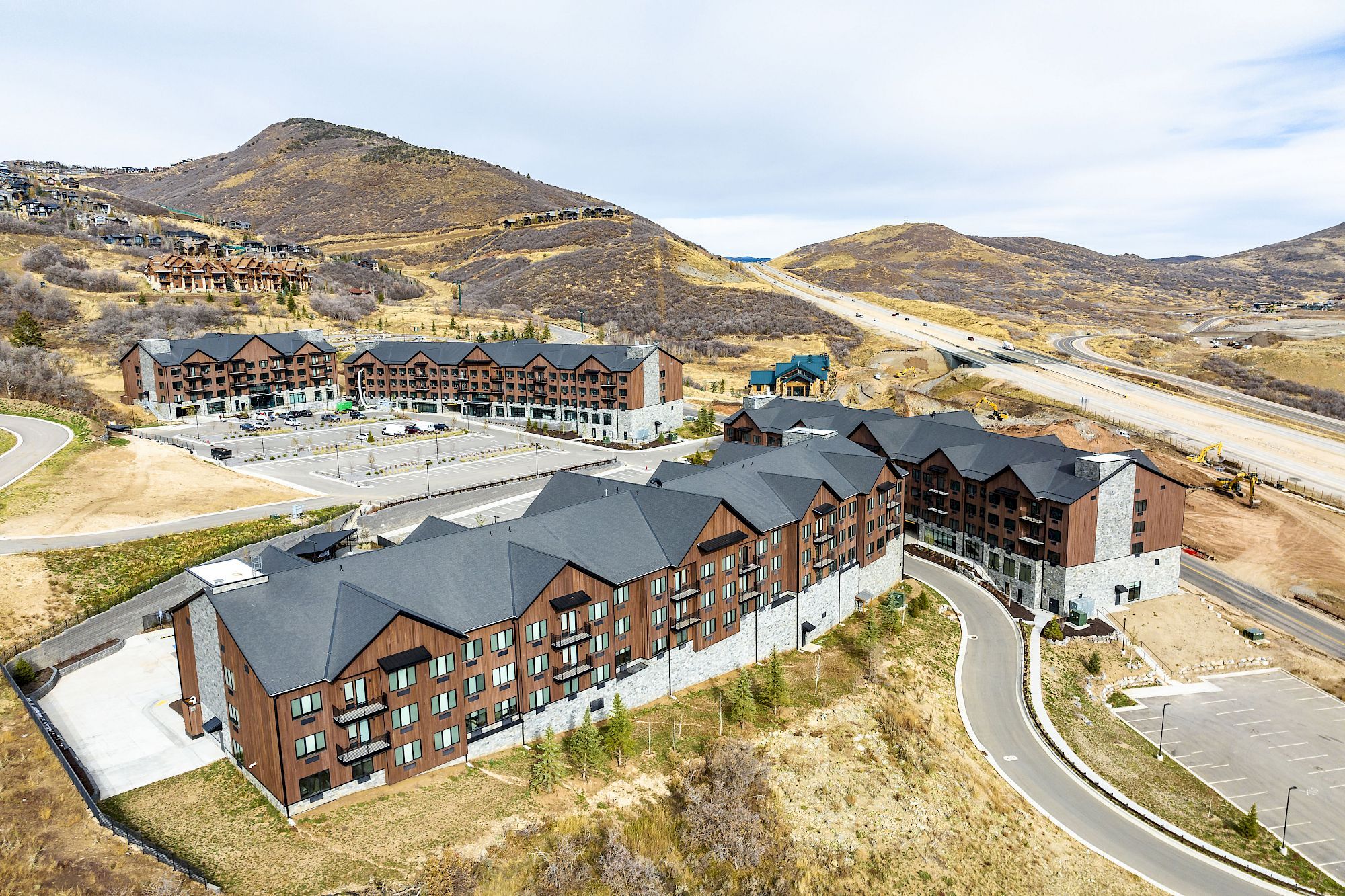 The image shows a complex of modern apartment buildings surrounded by hilly terrain, under a cloudy sky, with a road curving nearby.