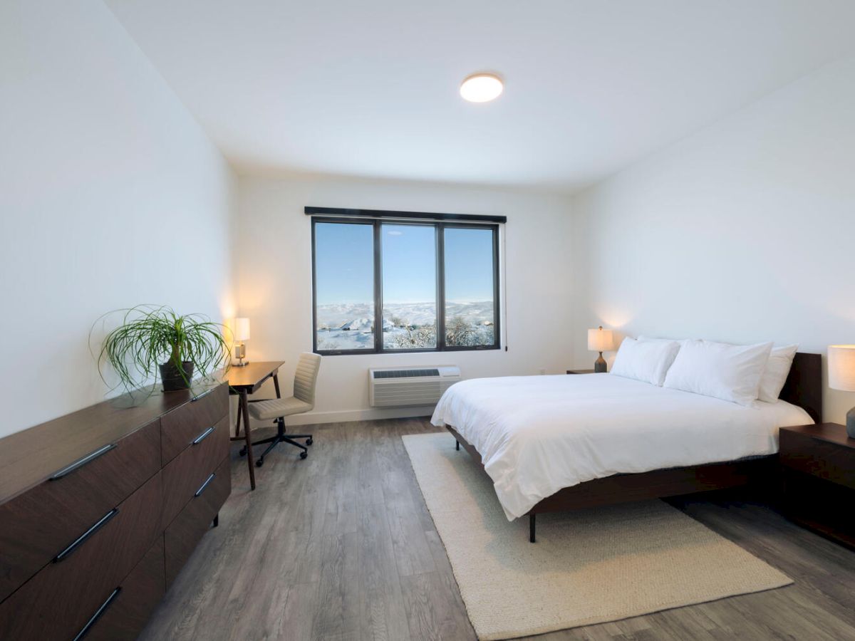 A modern bedroom with a large bed, two nightstands with lamps, a dresser with a plant, and a desk with a chair near a window.