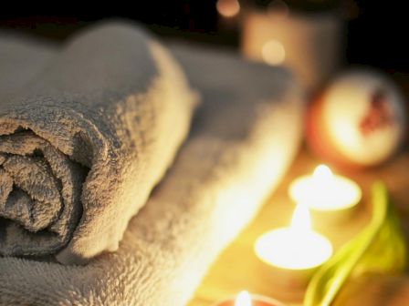 The image shows rolled towels, lit candles, and a purple tulip, creating a relaxing spa-like atmosphere on a wooden surface.
