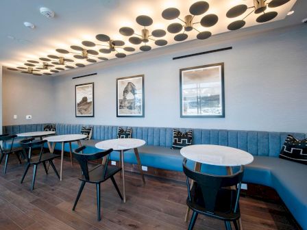 A modern seating area with round tables, black chairs, blue bench seating, decorative pillows, framed artwork on the wall, and unique ceiling lights.