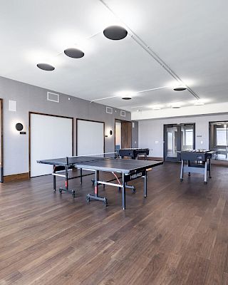 An empty game room with ping pong tables, arcade machines, and a seating area in the background, all on a wooden floor.