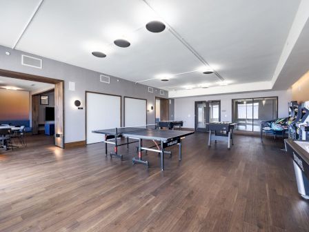 An empty game room with ping pong tables, arcade machines, and a seating area in the background, all on a wooden floor.