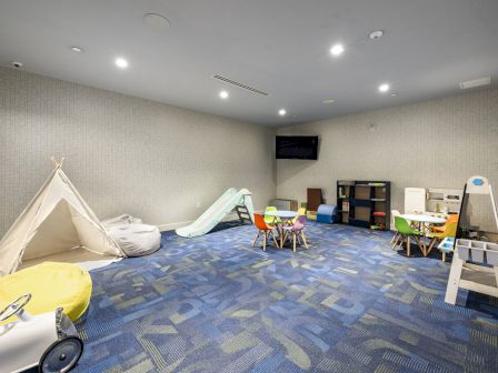 A children's playroom with a small tent, play slide, tables, chairs, toy shelves, and a TV on the wall. The floor is carpeted in blue.