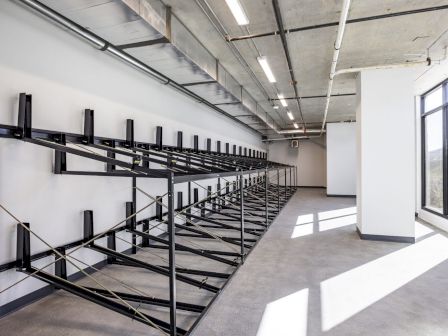 This image shows an empty industrial-style room with multiple rows of black metal racks, likely for storage, and large windows on one side.