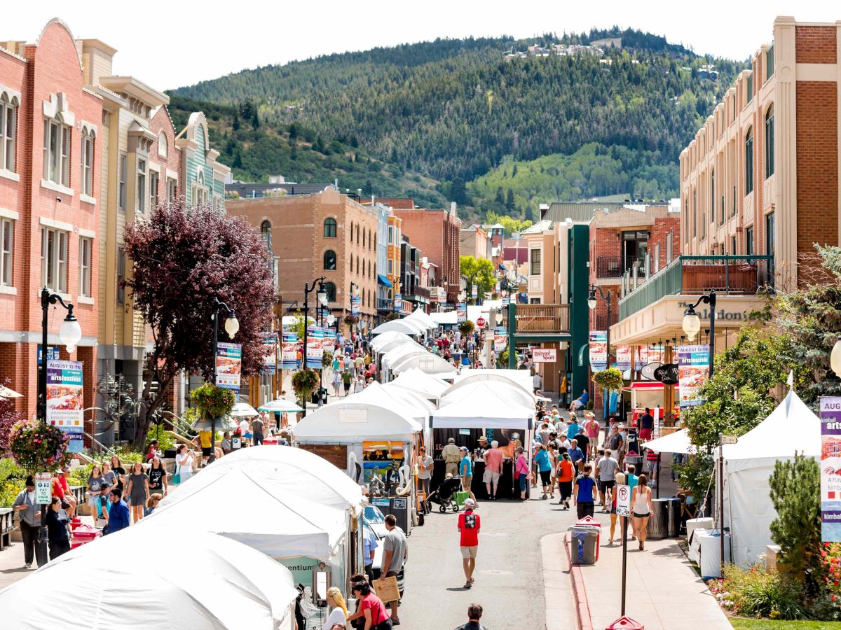 A bustling street fair in a sunny town, tents line the sidewalks, people stroll, and colorful storefronts hug the boardwalk with hills in the background.