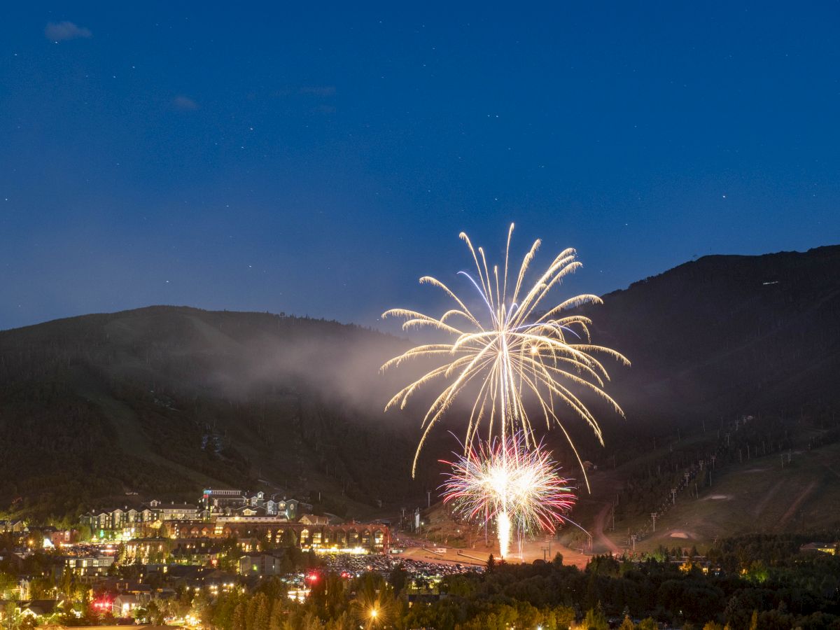 Fireworks bursting over a town at night, with mountains in the background and a faint cloud above the hills.