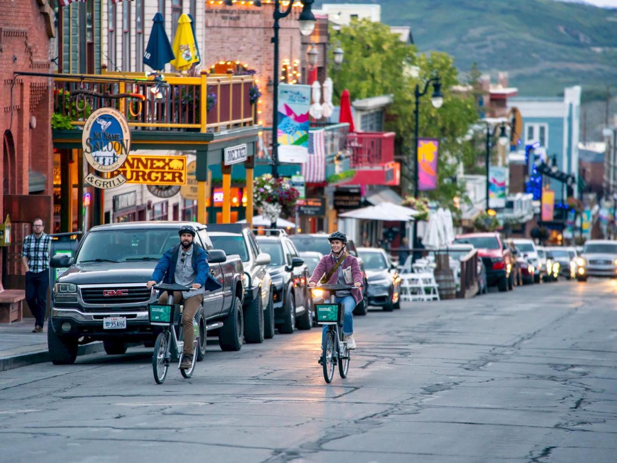 Street scene in a lively downtown: cars, bicycles, pedestrians, colorful storefronts, flags, mountains in the background.