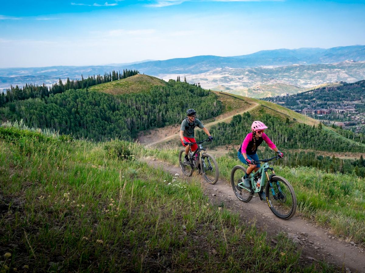 Two people mountain biking along a winding dirt trail on a grassy hillside, with a scenic valley and distant mountains in view.