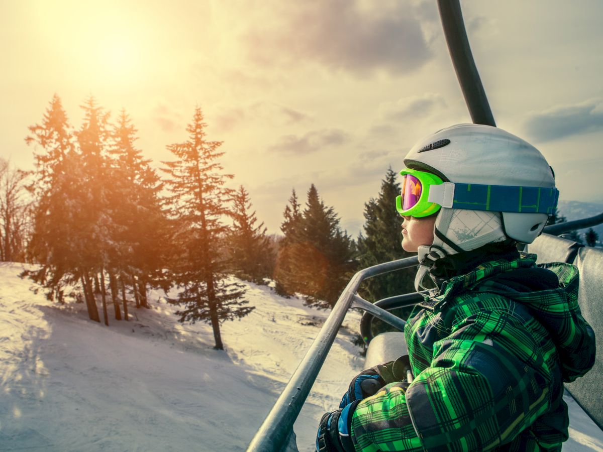 A child in ski gear is sitting on a ski lift, looking at a snowy landscape with sunlit trees in the background.