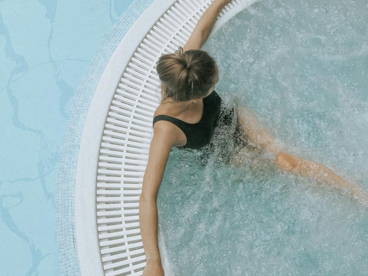 A person wearing a black swimsuit relaxes in a circular pool or hot tub with clear water, with their arms resting on the pool edge.