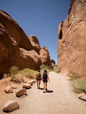 Two people are hiking through a narrow path surrounded by large red rock formations under a clear blue sky.