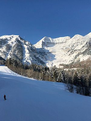 A lone skier descends a snowy mountain slope framed by picturesque, snow-covered peaks beneath a clear blue sky.