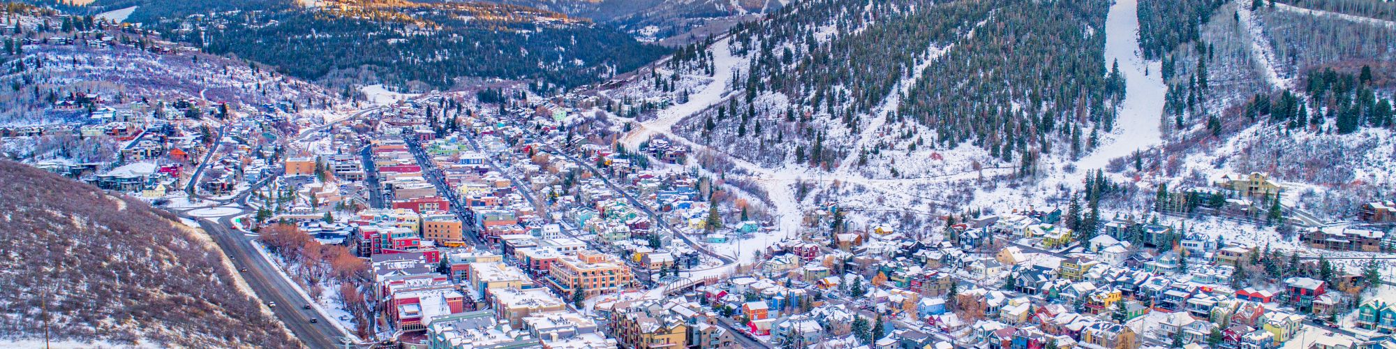 Aerial view of a snowy town nestled in the mountains, showcasing colorful buildings and ski slopes under a sunset sky.