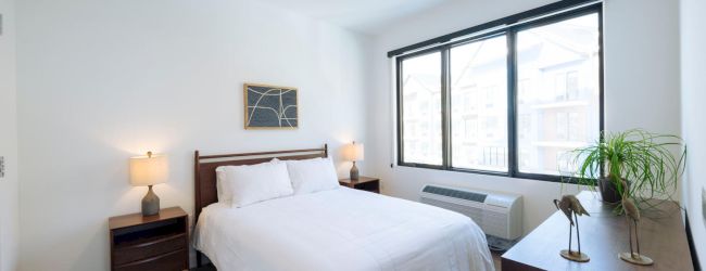 A modern bedroom with a bed, two bedside tables with lamps, a dresser with decor, and a large window. Light and minimalistic design.