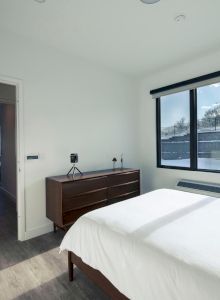 A minimalist bedroom with a bed, dresser, large window, and an adjoining room visible through an open door.