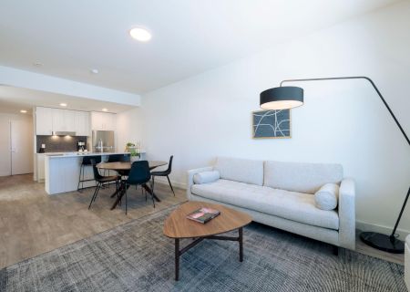 The image shows a modern living area with a sofa, coffee table, floor lamp, and a dining space leading to a kitchen with stools.