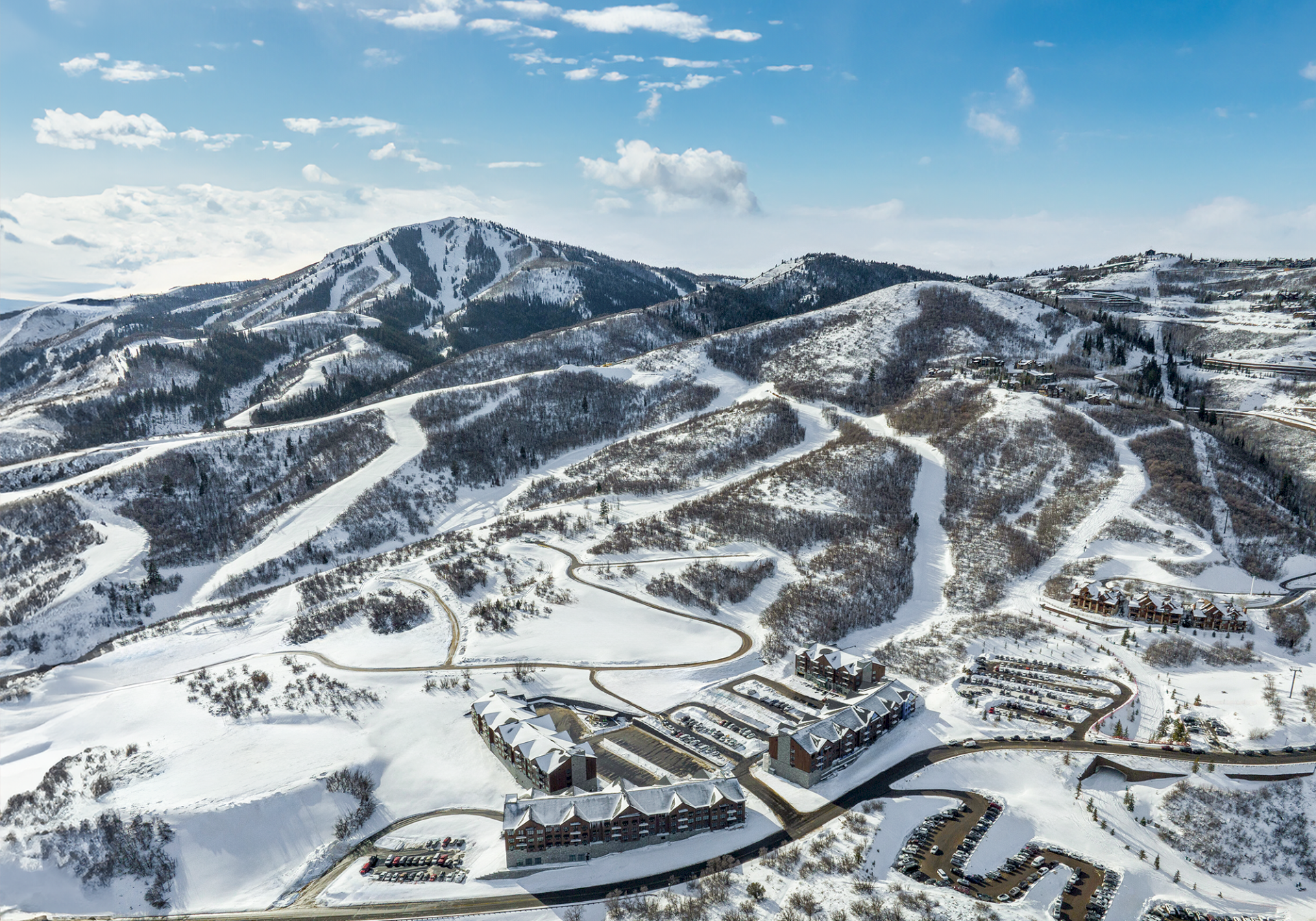 Snowy mountain resort with winding roads, ski lifts, and clustered buildings at base, nestled under a bright blue sky.