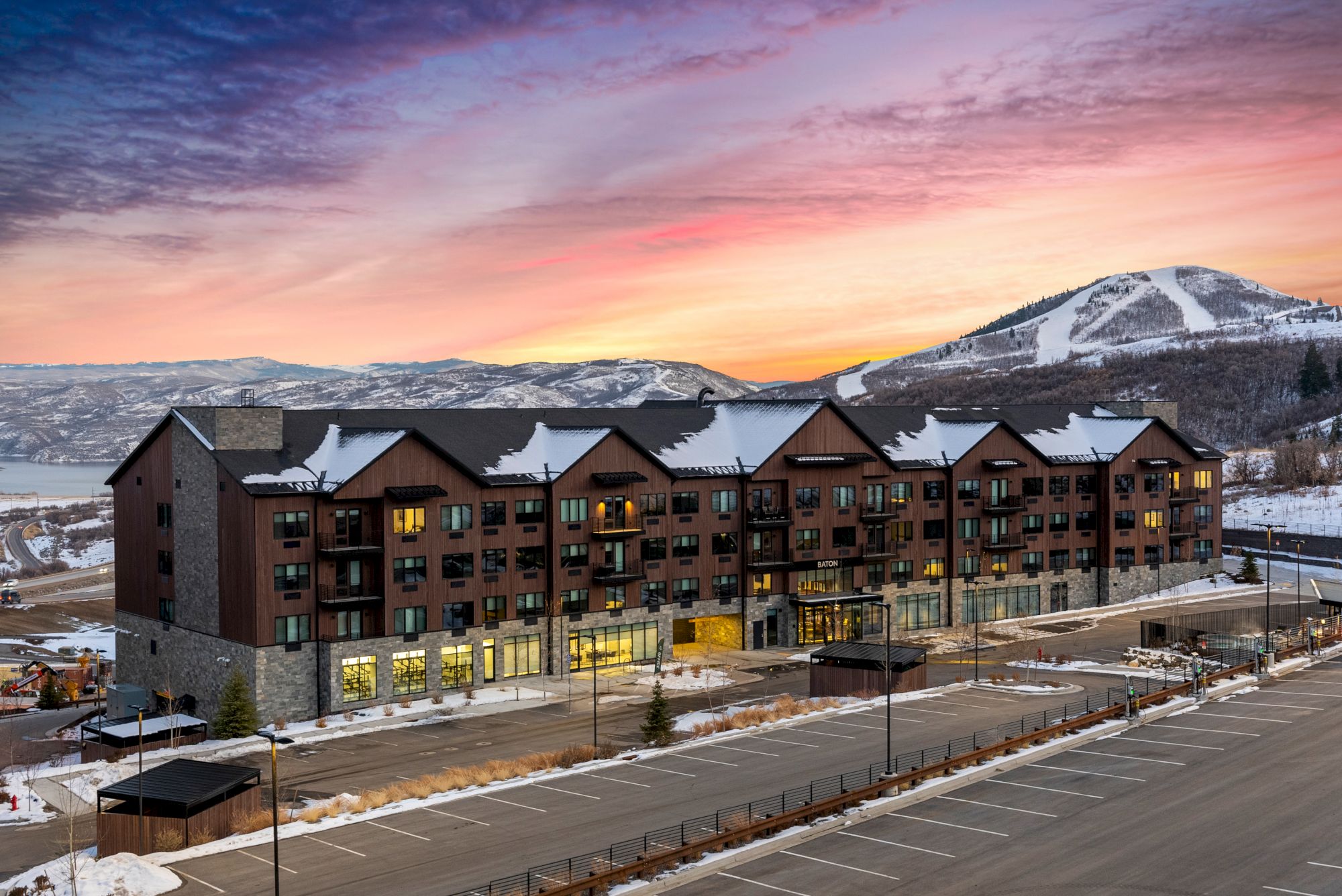 A large lodge-style building with a snowy mountain backdrop at sunset. The scene includes a parking lot and a serene winter landscape.