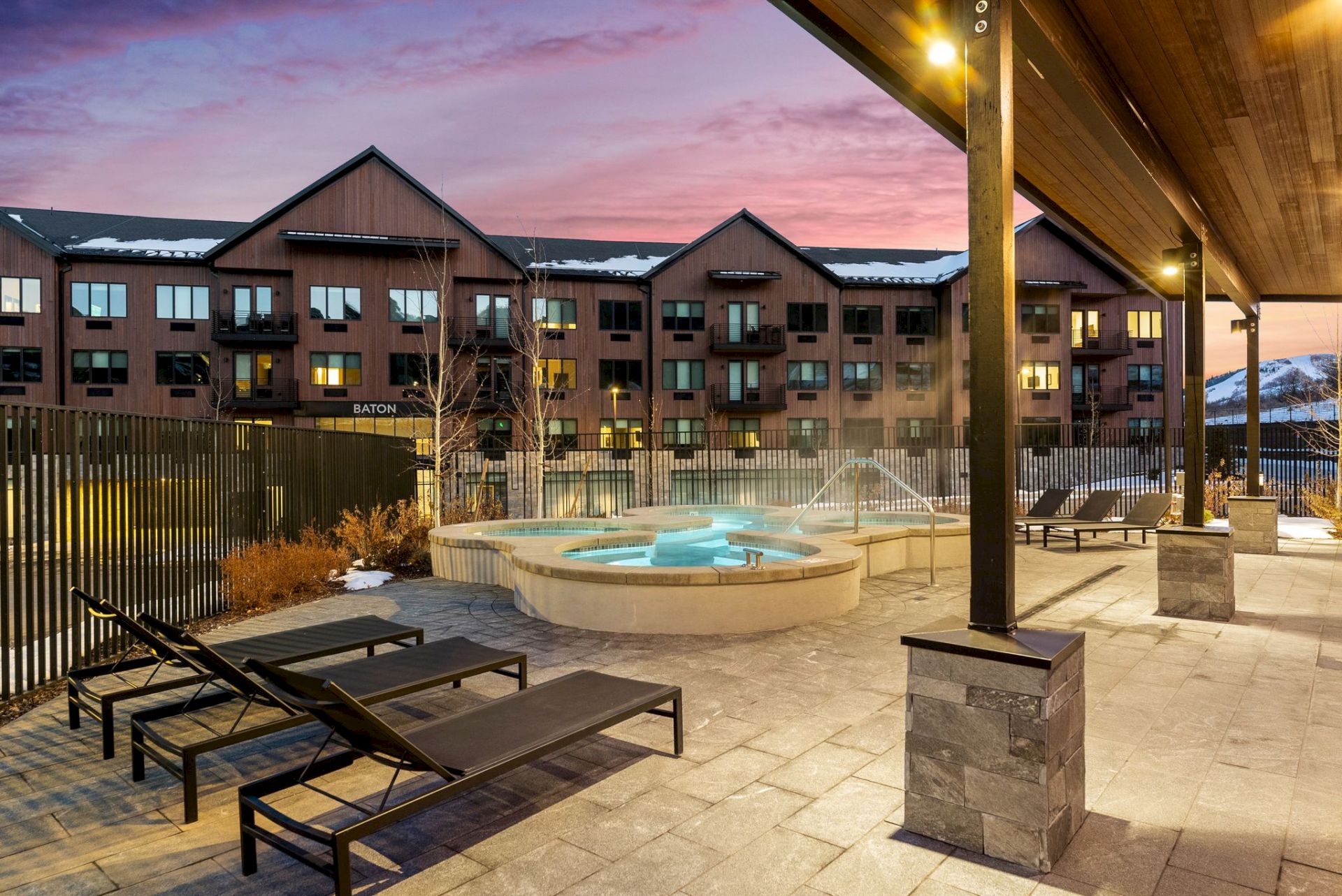 A modern hotel courtyard with a circular hot tub, lounge chairs, and a covered walkway, set against a sunset sky.