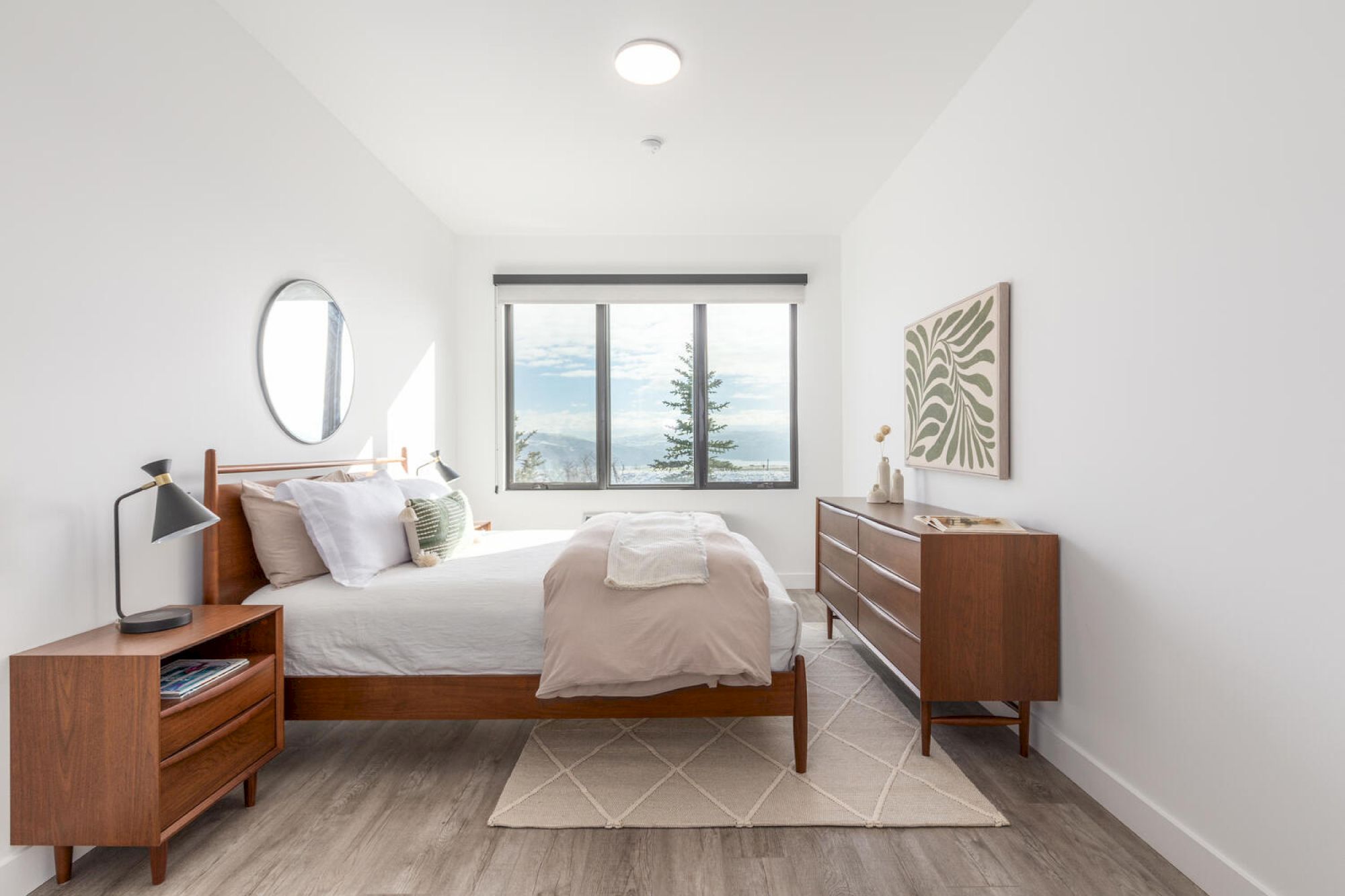 The image shows a minimalist bedroom with a wooden bed, nightstand, dresser, a circular mirror, and a window with a view outside.