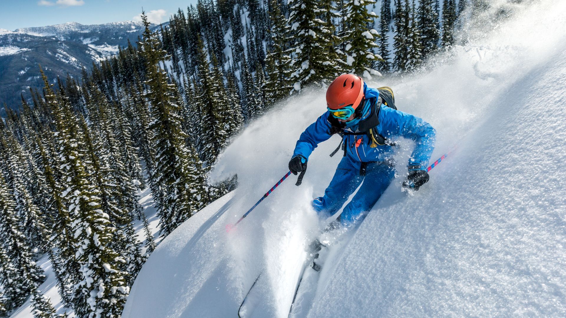 A skier in a blue suit and red helmet carving down a snow-covered slope, spraying powder with pine trees and mountains in the background.
