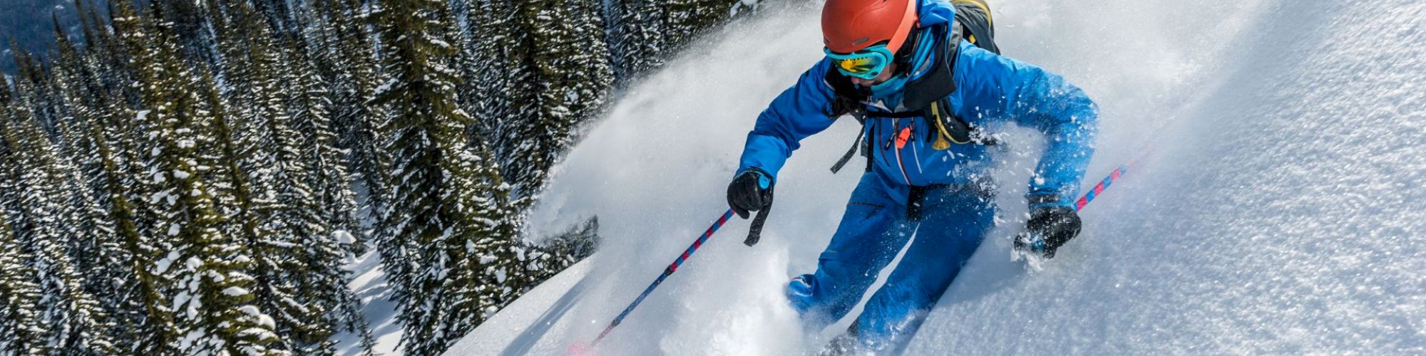 A skier in a blue suit and red helmet carving down a snow-covered slope, spraying powder with pine trees and mountains in the background.