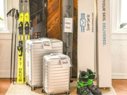 A cozy corner shows travel gear: skis, a couple of silver suitcases, a backpack, and a pair of green-and-black boots, ready for a trip.