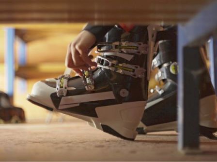 A person lacing up a mid-height ski or snowboard boot under a bench, getting ready to ride.