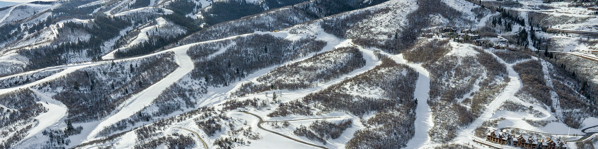 Snowy mountain resort with winding roads, ski trails, and clustered buildings below, all under a clear blue sky, inviting winter fun.