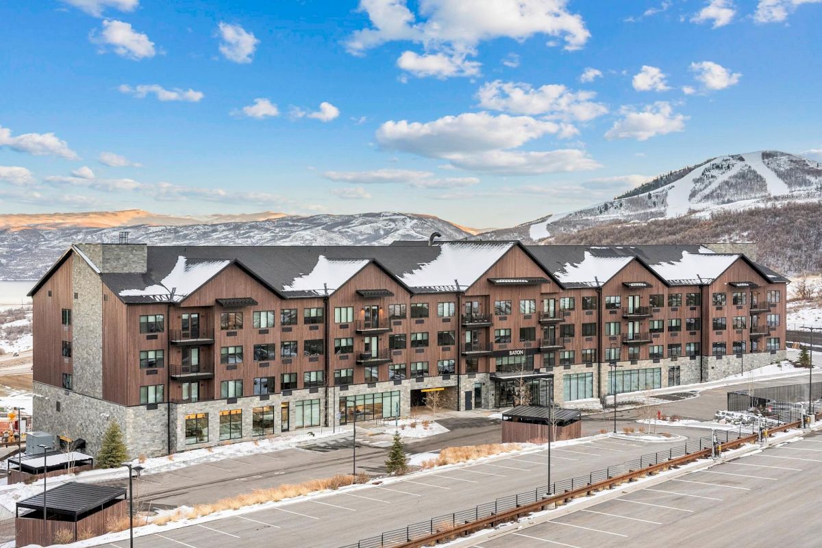 A large, multi-story building with a brown and stone exterior, snow-covered roof, and balconies, set against a backdrop of snow-capped mountains and a blue sky with clouds.