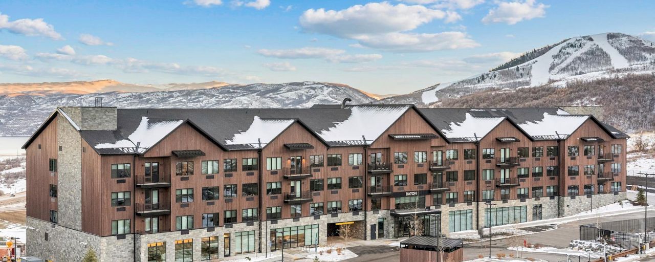 A multi-story hotel building with a brown and stone facade, snow-covered roof, and balconies, set against a backdrop of snowy mountains and a blue sky with clouds.