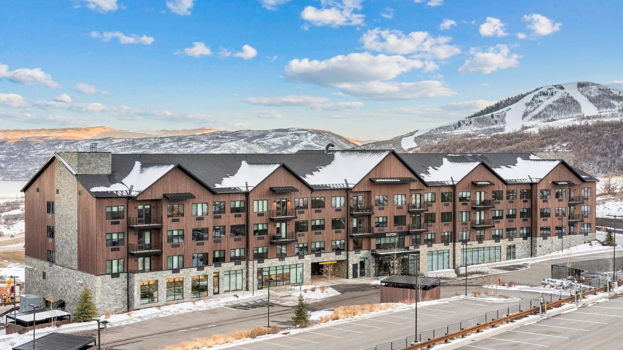 A large, multi-story building with a brown and stone exterior, snow-covered roof, and balconies, set against a backdrop of snow-capped mountains under a blue sky with clouds.