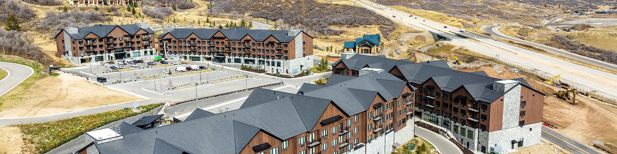 A large wooden hotel/resort complex sits in a mountain valley, with winding roads and hills under a cloudy sky.