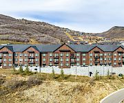 A row of modern brick townhouses with dark roofs sits along a curved road, set in a dry, hilly landscape under a blue sky.