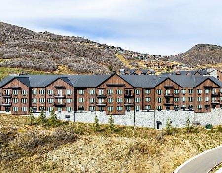 A row of modern brick townhouses with dark roofs sits along a curved road, set in a dry, hilly landscape under a blue sky.