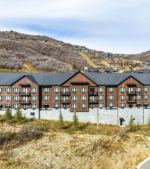 A row of modern brick townhouses with dark roofs sits along a curved road, set in a dry, hilly landscape under a blue sky.