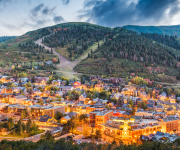 A small town nestled at the foothills, with colorful buildings lit warmly, surrounded by forested slopes and a cloudy mountain backdrop.
