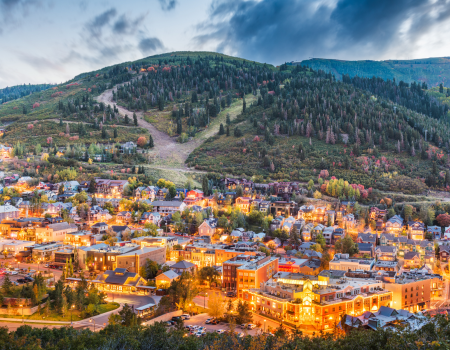 A small town nestled at the foothills, with colorful buildings lit warmly, surrounded by forested slopes and a cloudy mountain backdrop.