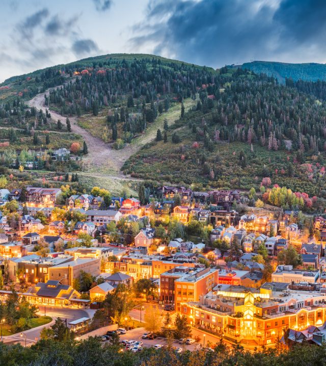 A small town nestled at the foothills, with colorful buildings lit warmly, surrounded by forested slopes and a cloudy mountain backdrop.