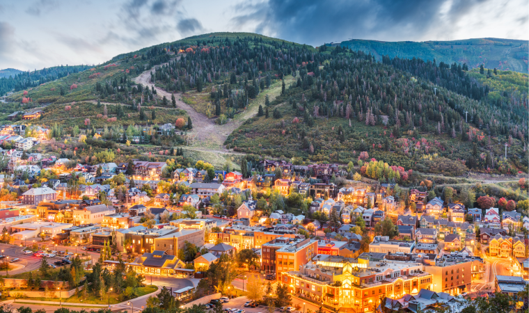 A small town nestled at the foothills, with colorful buildings lit warmly, surrounded by forested slopes and a cloudy mountain backdrop.